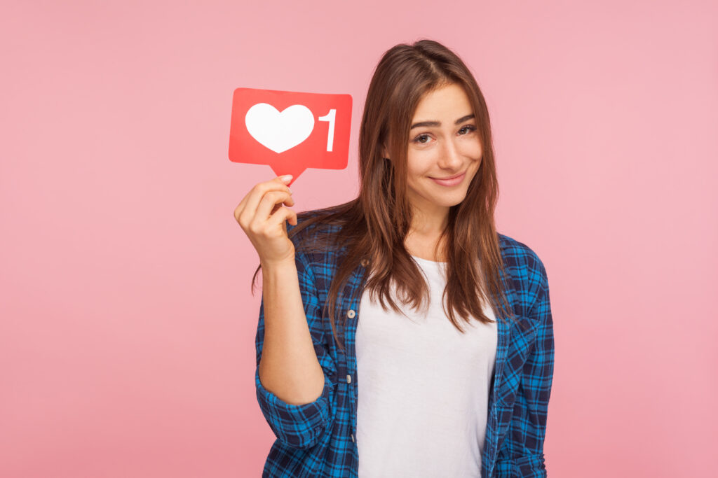 Ritratto di una ragazza carina e positiva con una camicia a scacchi che sorride scherzosamente e tiene in mano l'icona dei social media, il pulsante del cuore per godersi contenuti interessanti e alla moda. Rappresenta la psicologia Marketing del beauty nel mondo dei social media