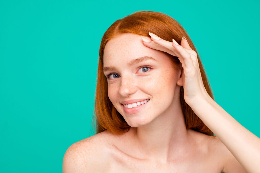 Ritratto ravvicinato di una ragazza dai capelli rossi affascinante, bella e naturale, nuda, con la pelle liscia, pura e lucida, che tocca la fronte, isolata su sfondo verde turchese. l'immagine vuole rappresentare l'utilizzo dei principi attivi su una pelle giovane e adolescenziale