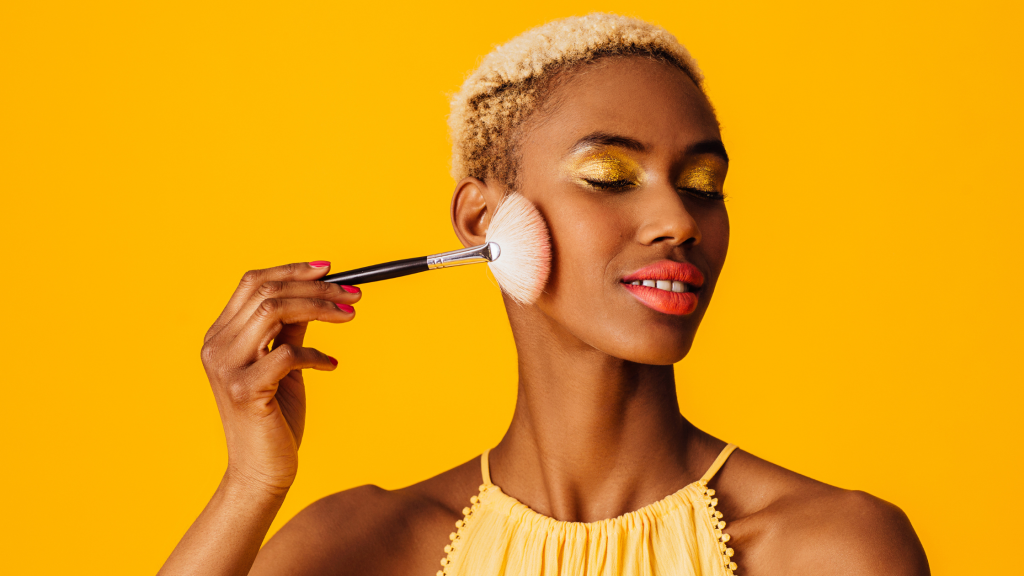 una donna su sfondo giallo che si trucca. Rappresenta l'immagine di cosa non deve mai mancare nel nostro beauty estivo