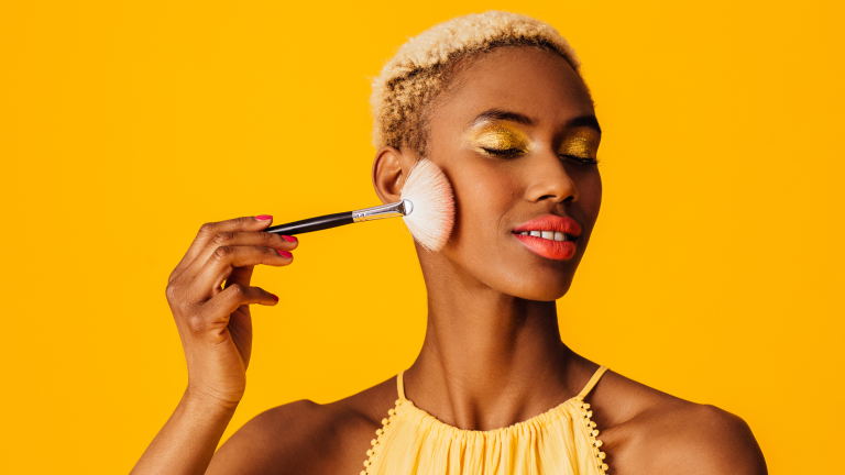 una donna su sfondo giallo che si trucca. Rappresenta l'immagine di cosa non deve mai mancare nel nostro beauty estivo