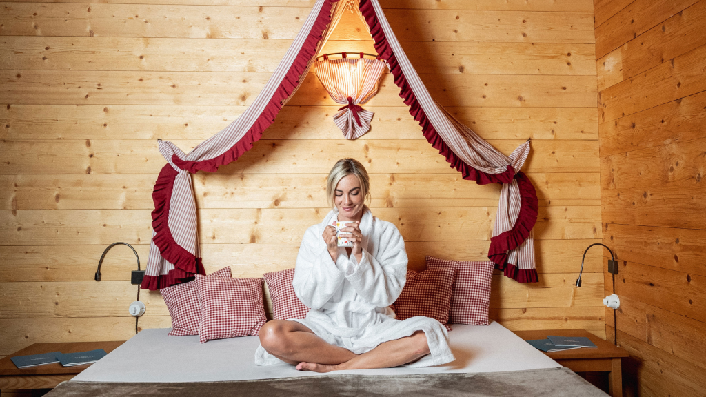 Donna sorridente in accappatoio bianco seduta a gambe incrociate sul letto in una stanza in stile alpino con pannelli di legno, mentre tiene tra le mani una tazza bianca. Il letto presenta lenzuola chiare e cuscini rossi e bianchi a quadri. Una tenda a baldacchino rossa e bianca pende sopra la testa, creando un'atmosfera accogliente e rilassante, tipica di una longevity spa o resort benessere.