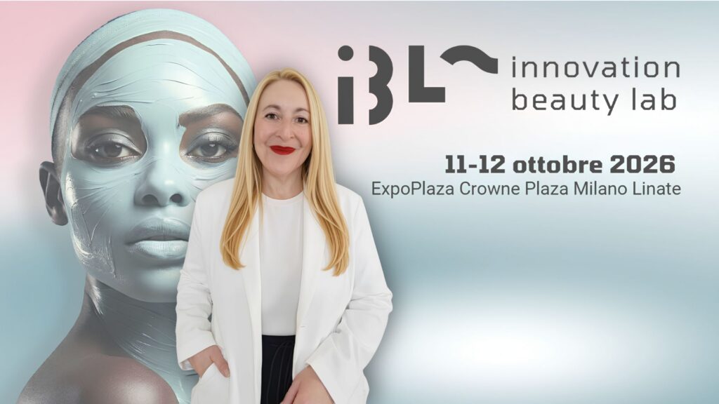 Una donna bionda con giacca e sottogiacca bianchi, pantaloni neri, tieni una mano in tasca e sosta, sorridente davanti al banner visual di IBL Innovation Beauty Lab 2026