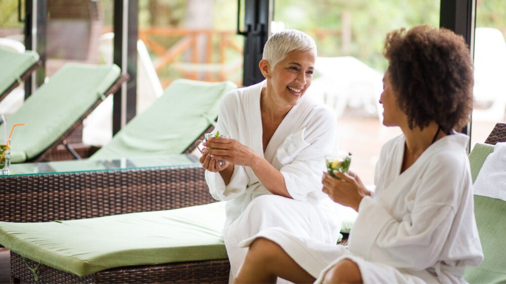 Una donna matura e bionda e una donna più giorvane di colore sorseggiano una tisana nella zona relax di una spa. L'ambiente è composto da grandi vetrate da cui si intravede il verdeggiare di un parco e i lettini sono color bianco e verde pistacchio chiaro.