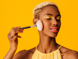 una donna su sfondo giallo che si trucca. Rappresenta l'immagine di cosa non deve mai mancare nel nostro beauty estivo