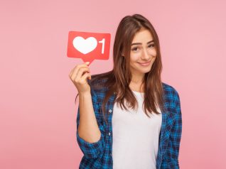 Ritratto di una ragazza carina e positiva con una camicia a scacchi che sorride scherzosamente e tiene in mano l'icona dei social media, il pulsante del cuore per godersi contenuti interessanti e alla moda. Rappresenta la psicologia Marketing del beauty nel mondo dei social media