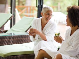 Una donna matura e bionda e una donna più giorvane di colore sorseggiano una tisana nella zona relax di una spa. L'ambiente è composto da grandi vetrate da cui si intravede il verdeggiare di un parco e i lettini sono color bianco e verde pistacchio chiaro.