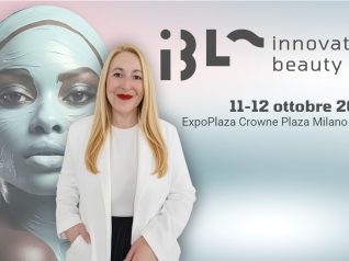 Una donna bionda con giacca e sottogiacca bianchi, pantaloni neri, tieni una mano in tasca e sosta, sorridente davanti al banner visual di IBL Innovation Beauty Lab 2026